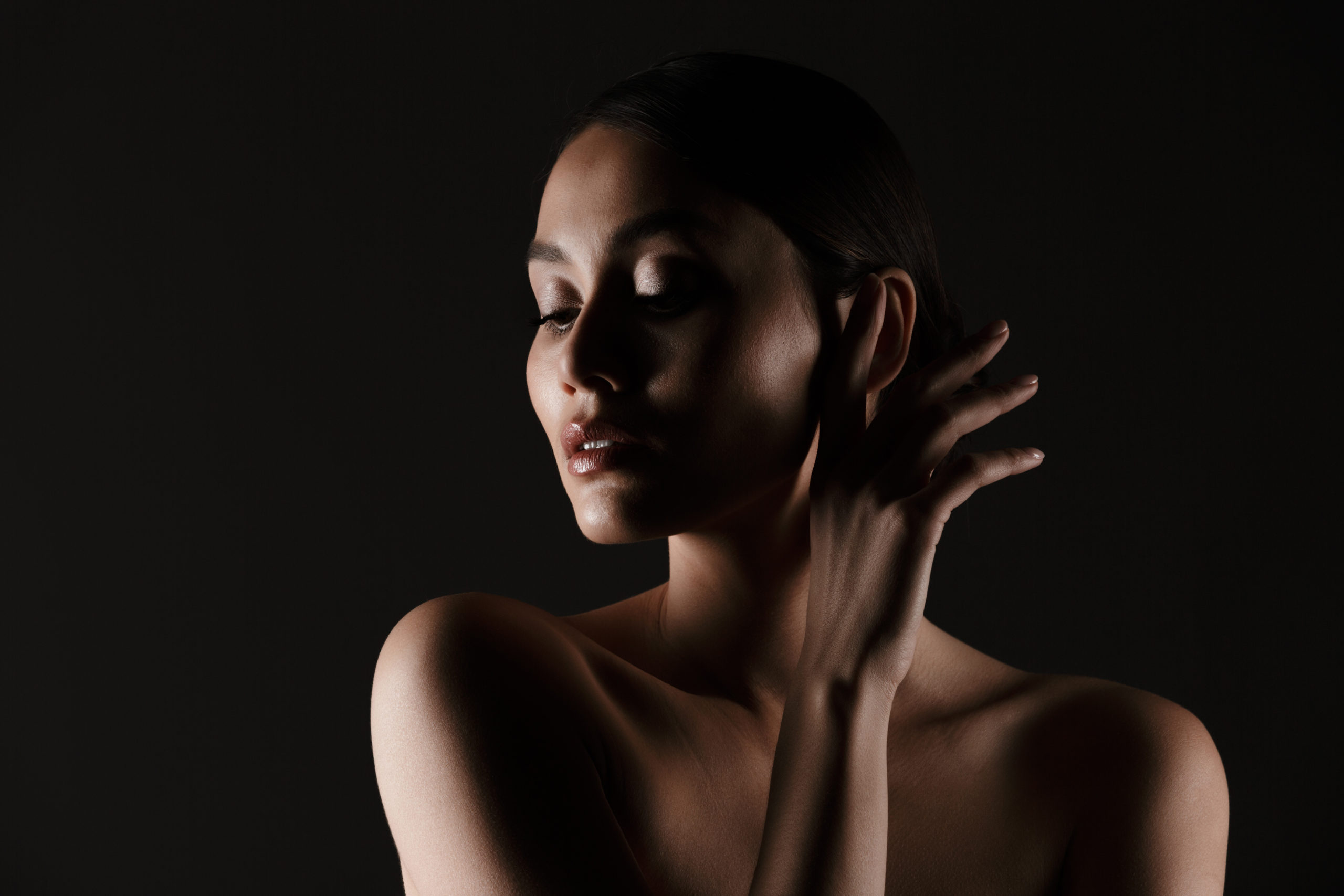 Portrait of feminine tender woman with sensual look posing at camera looking aside in low lights, isolated over black background